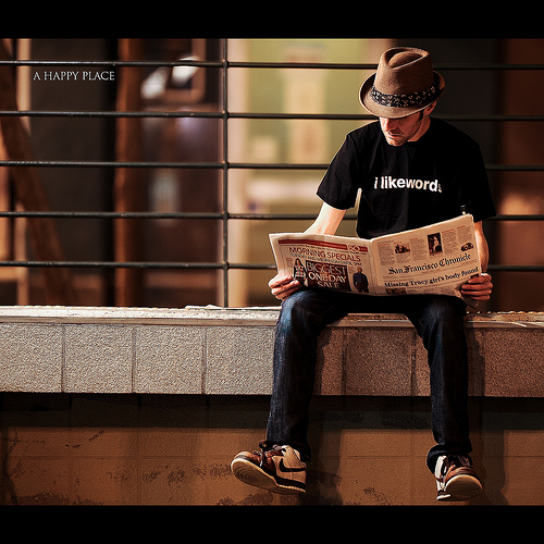 hipster man in trilby hat with t-shirt that says "i like words" reading a newspaper while sitting on a loading dock.